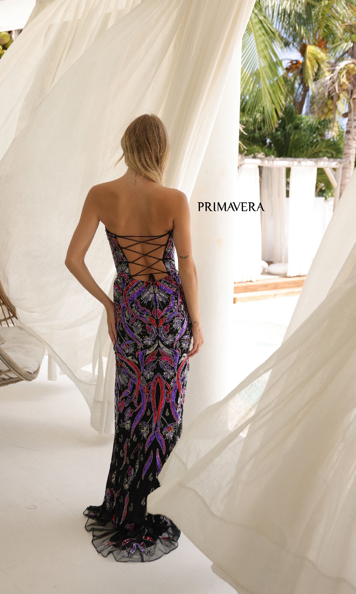 A woman with blonde hair wears the Primavera 4387 long prom dress—a black, backless gown with a sweetheart neckline and colorful pattern—standing outdoors by white curtains and columns.
